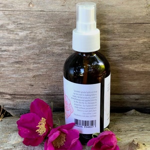 May include: A brown glass spray bottle with a white pump and label, next to three vibrant pink rose blossoms. The label text describes the product as a cosmetic grade hydrosol. The bottle is on a wooden surface.