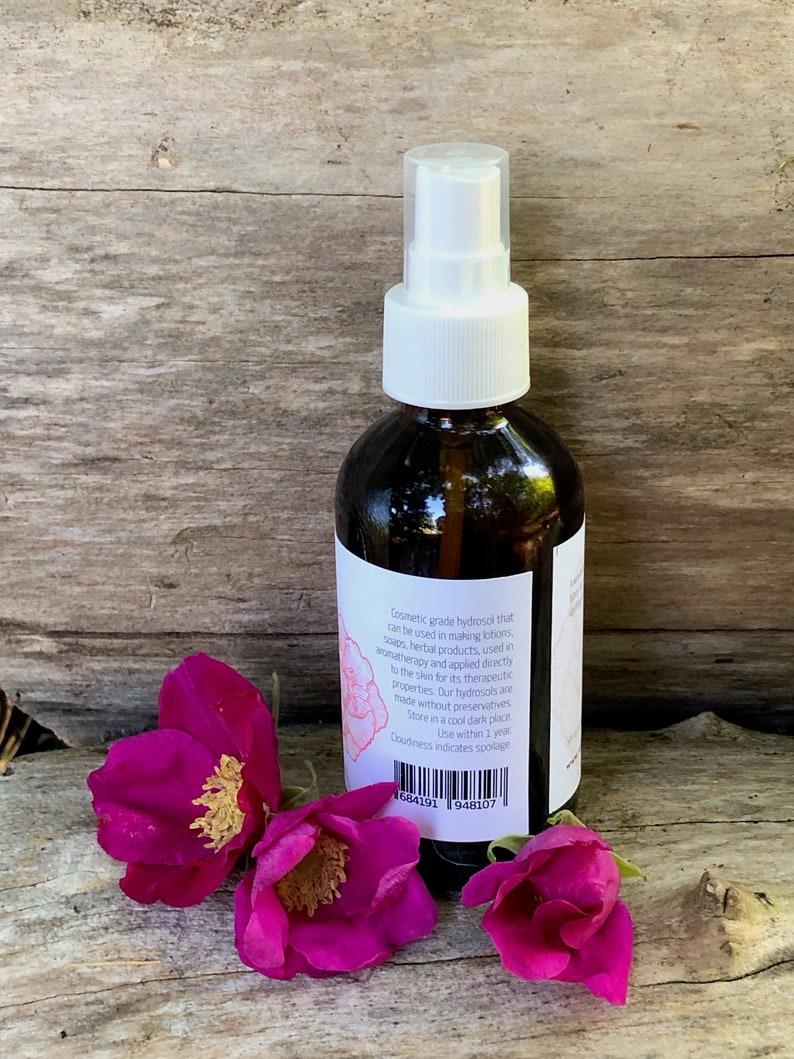 May include: A brown glass spray bottle with a white pump and label, next to three vibrant pink rose blossoms. The label text describes the product as a cosmetic grade hydrosol. The bottle is on a wooden surface.