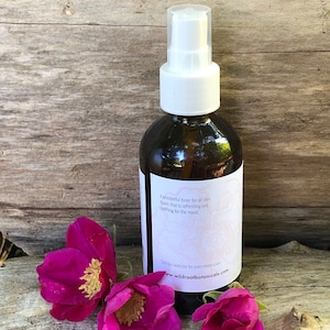 May include: A dark brown glass spray bottle with a white top and a label. The label reads "A wonderful toner for all skin types that is refreshing and uplifting for the mood." Three pink flowers are in front of the bottle.