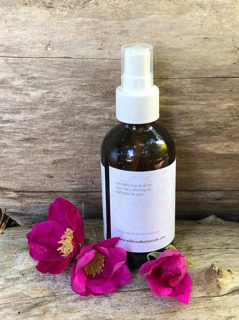 May include: A dark brown glass spray bottle with a white top and a label. The label reads "A wonderful toner for all skin types that is refreshing and uplifting for the mood." Three pink flowers are in front of the bottle.