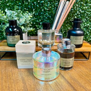 May include: Five glass bottles of reed diffusers with different scents. The bottles are on a wooden shelf with a green background. The scents are "Peach Cottage", "Mango &  Hibiscus", "Amber & Bay Leaf", "Black Coral & Smoke", and "Fig &  Oakmoss".