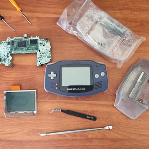 Könnte beinhalten: Ein zerlegter Game Boy Advance SP mit einer violetten Hülle, einer durchsichtigen Hülle, einem Motherboard, einem Bildschirm und Werkzeugen zur Reparatur.