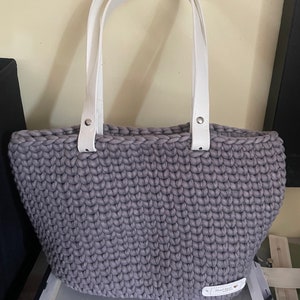Peut inclure: Un sac cabas en crochet gris avec des anses en cuir blanc. Le sac a une petite étiquette en cuir blanc avec le texte "Hand Made" et un cœur.