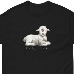 Puede incluir: Camiseta negra con una ilustración detallada de un cordero blanco con una marca roja en forma de diamante en la frente. El texto "Ruby Lamb" está impreso debajo del cordero en una fuente estilizada. La camiseta está hecha de un material suave.