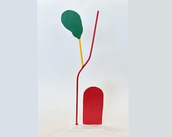 Op de afbeelding: Abstracte sculptuur met een groene bladvorm, een gele steel en een rode gebogen lijn en rechthoekige vorm. De sculptuur is gemonteerd op een heldere basis, met een witte achtergrond.