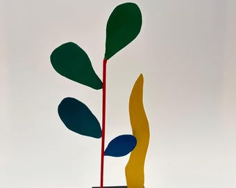 Op de afbeelding: Een kleurrijke abstracte metalen sculptuur met een rode stengel, groene en blauwe bladvormen en een gele, vlamachtige vorm. De sculptuur is gemonteerd op een zwarte vierkante basis.