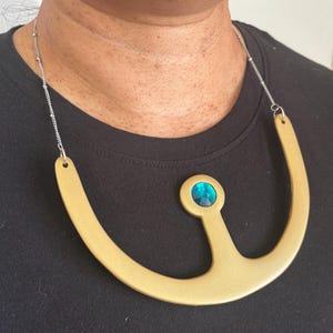 Puede incluir: Un collar dorado con un colgante grande y una cadena plateada. El colgante tiene un diseño curvo con una piedra preciosa central redonda de color turquesa. El collar se lleva sobre una camiseta negra.