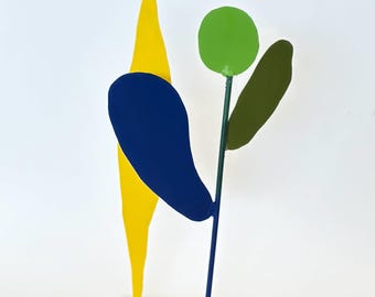 Op de afbeelding: Abstracte metalen sculptuur met een geel, blauw en groen kleurenpalet. De sculptuur heeft een gele, puntige vorm, een blauwe druppelvorm en een groene cirkel en olijfgroene bladvorm op een groene stengel. Het staat op een witte basis.