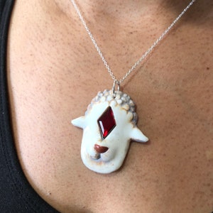 Puede incluir: Un colgante de cerámica blanca en forma de oveja con una piedra preciosa roja en forma de diamante para el ojo, colgado de una cadena plateada.