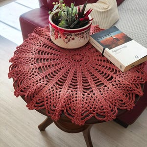 May include: A circular, terracotta-coloured crochet doily on a dark wooden table. A small potted succulent plant with red detailing and a book titled "Agustos Işığı" are on the table. The doily features an intricate, openwork pattern.