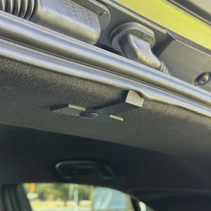 May include: A detailed shot of a car's interior roof lining. The dark grey panel features a textured finish. A black plastic component with a geometric shape is affixed to the panel. A black rubber tube is also visible.