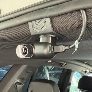 May include: A black dash camera mounted inside a vehicle. The camera is attached to the headliner with a black bracket. A USB cable is plugged into the camera. The camera has the text "V-OUT" printed on it.