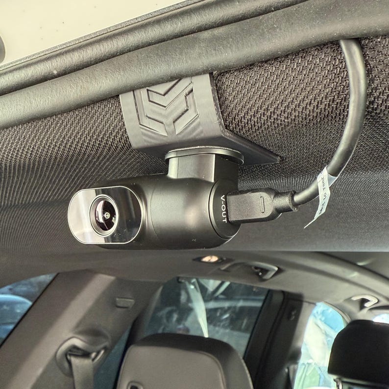 May include: A black dash camera mounted inside a vehicle. The camera is attached to the headliner with a black bracket. A USB cable is plugged into the camera. The camera has the text "V-OUT" printed on it.