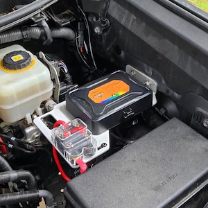 May include: A black electrical component with an orange and rainbow label is mounted inside a vehicle's engine compartment. A clear fuse holder with red wiring is also visible, along with other mechanical parts.