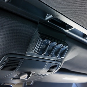 May include: A black switch panel with four toggle switches mounted in a car's overhead console. The switches have a black plastic housing and a silver metal button.