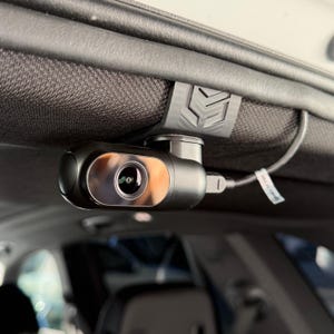 Op de afbeelding: Een zwarte dashcam gemonteerd in een voertuig. De camera heeft een zilveren lens en is aangesloten op een kabel. Hij is met een zwarte clip aan de hemelbekleding bevestigd.