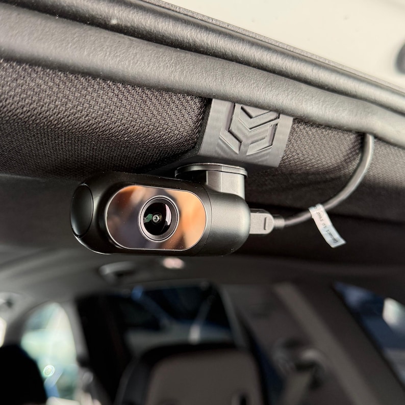 May include: A black dash camera mounted inside a vehicle. The camera has a silver lens and is connected to a cable. It is attached to the headliner with a black clip.