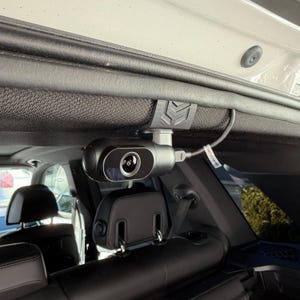 May include: A black car dash camera mounted on the interior roof of a vehicle. The camera is cylindrical with a lens and is connected to a wire. The camera is attached to a black bracket.
