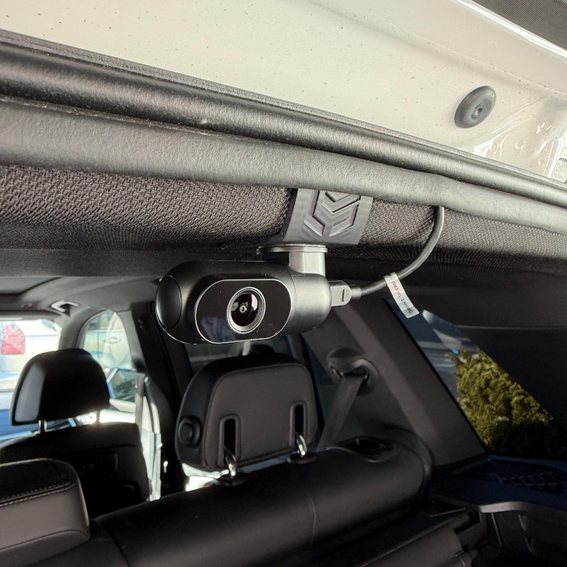 May include: A black car dash camera mounted on the interior roof of a vehicle. The camera is cylindrical with a lens and is connected to a wire. The camera is attached to a black bracket.