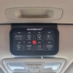 Puede incluir: Un panel de interruptores AUXBEAM negro con 8 botones para controlar las luces. Los botones están etiquetados: Barra de luz, Capó, Inundación, Luces antiniebla, Lateral, Roca, Estroboscópico y DRL. El panel tiene un interruptor de encendido/apagado rojo.