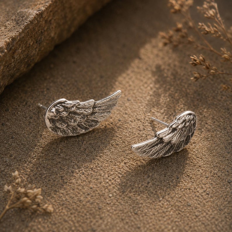 May include: A pair of silver wing-shaped stud earrings. The earrings feature detailed feather textures and are presented on a textured, neutral-toned surface. The earrings are designed to be worn on the ear.
