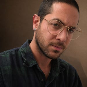 May include: A person wearing gold-rimmed spectacles and a dark green and blue checked shirt. The person has a short haircut and a small earring. The background is a solid brown colour.