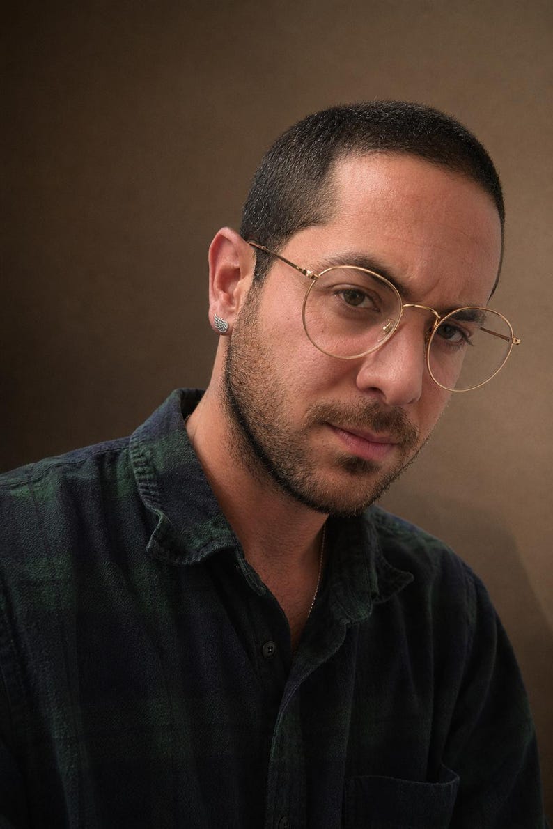 May include: A person wearing gold-rimmed spectacles and a dark green and blue checked shirt. The person has a short haircut and a small earring. The background is a solid brown colour.