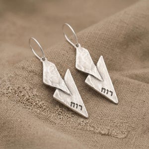May include: A pair of silver-toned drop earrings featuring a textured, geometric, layered design. Each earring has a hook-style closure and displays the Hebrew word "רוח" engraved in black lettering on the lower triangular section of the metal.