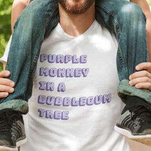 Op de afbeelding: Een wit T-shirt met de tekst "Purple Monkey in a Bubblegum Tree" in paars met een 3D-effect.