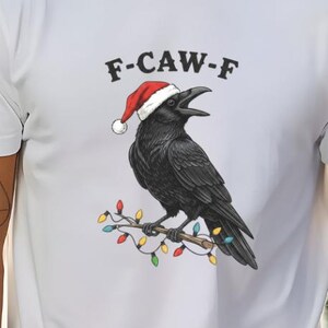 Puede incluir: Camiseta blanca con un cuervo negro con un gorro de Papá Noel y posado en una rama adornada con luces navideñas de colores. El texto "F-CAW-F" está encima del cuervo.
