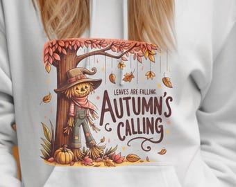 May include: White hoodie with a graphic of a scarecrow, tree, and the text "Autumn's Calling." The design features fall colors like orange, brown, and yellow. The person in the image is wearing a white beanie and blue jeans.