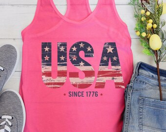 May include: A bright pink racerback tank top featuring the text USA in a distressed navy blue, white, and red flag pattern. Below the text, the words SINCE 1776 are printed in navy blue, flanked by two small navy blue stars on each side.
