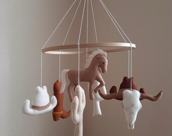 May include: A hanging mobile featuring felt figures suspended from a circular wooden frame. The figures include a white cowboy hat, a brown boot, a tan horse, a beige cactus, and a white steer skull with brown horns, all attached by white strings.