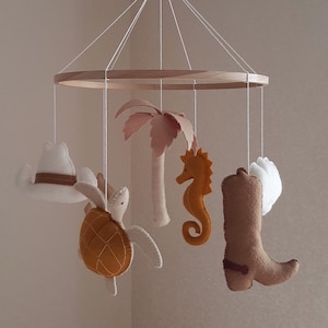 May include: A hanging mobile featuring a circular wooden frame with white strings suspending felt shapes including a white cowboy hat, a brown sea turtle, a light pink palm tree, an orange seahorse, and a brown cowboy boot with a dark brown spur.