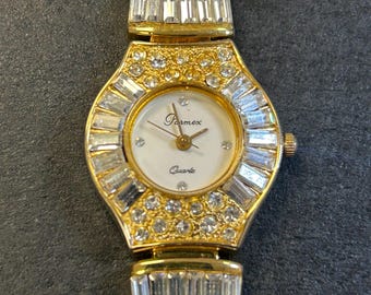 Può includere: Un orologio da polso dorato con quadrante bianco e il marchio "Parmex". L'orologio ha un movimento al quarzo ed è adornato da numerosi cristalli trasparenti e scintillanti. Il cinturino e la lunetta sono impreziositi da cristalli rettangolari e rotondi.