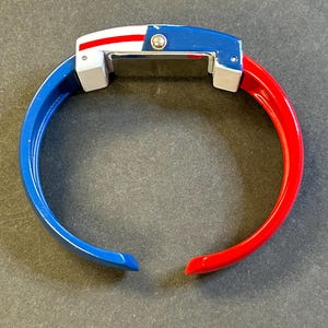 May include: A vibrant, open-ended bangle watch with a patriotic color scheme. The watch features a blue top with a white and red stripe design, and the bangle is split into blue and red sections. The watch has a rectangular face.