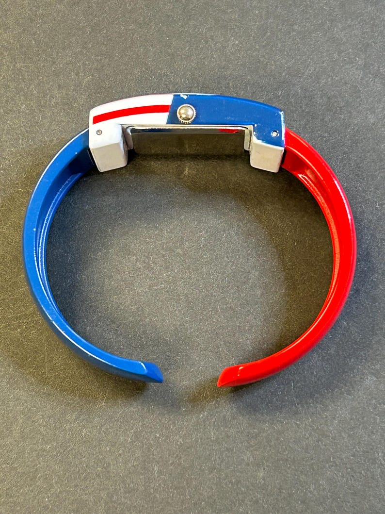 May include: A vibrant, open-ended bangle watch with a patriotic color scheme. The watch features a blue top with a white and red stripe design, and the bangle is split into blue and red sections. The watch has a rectangular face.