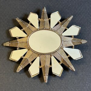 May include: A gold-toned starburst brooch with a white mirror in the center. The brooch has a geometric design with pointed rays.