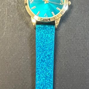 May include: A teal glitter watch with a gold-colored case and a matching teal glitter band. The watch face is teal with gold-colored hands and hour markers. The band has a buckle closure.