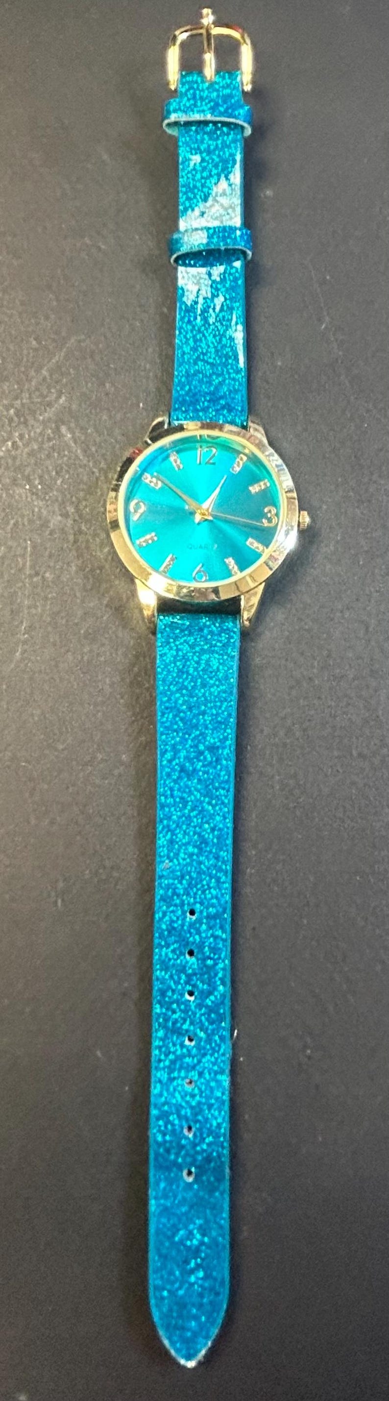 May include: A teal glitter watch with a gold-colored case and a matching teal glitter band. The watch face is teal with gold-colored hands and hour markers. The band has a buckle closure.