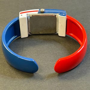 May include: A colorful bangle watch with a rectangular face. The watch features a blue, white, and red color scheme. The band is split into blue and red sections. The watch face is silver.