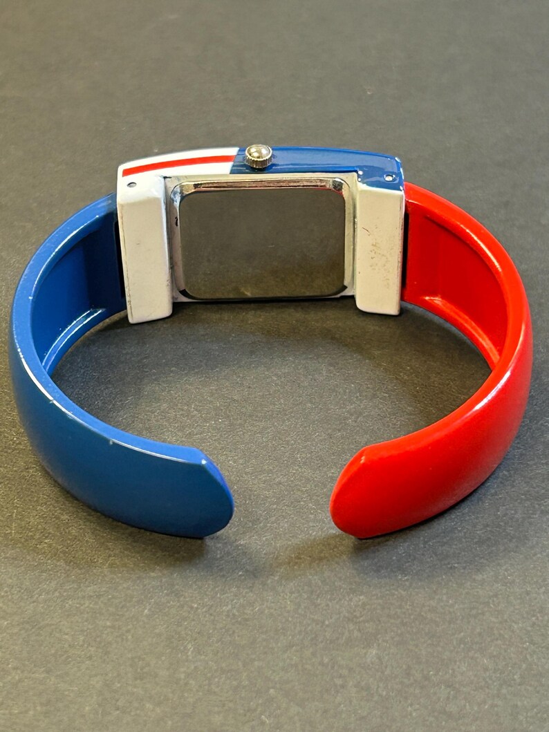 May include: A colorful bangle watch with a rectangular face. The watch features a blue, white, and red color scheme. The band is split into blue and red sections. The watch face is silver.