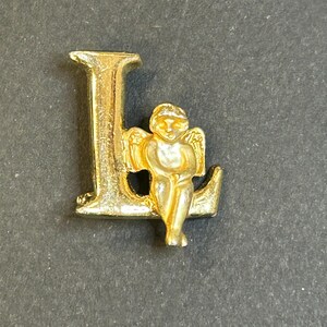 May include: Gold-coloured letter L charm with a small angel figurine sitting on the top of the letter.