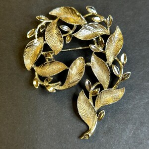 Könnte beinhalten: Goldfarbene Metallbrosche im Blätterdesign. Die Brosche zeigt eine kreisförmige Anordnung detaillierter Blätter und Stiele mit strukturierter Oberfläche. Die Blätter variieren in der Größe und sind in einem fließenden, organischen Muster angeordnet.