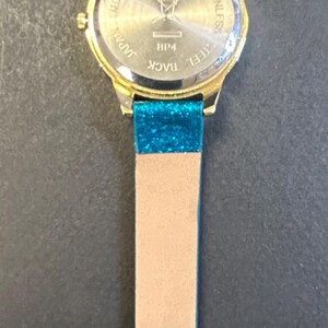 May include: Gold-toned wristwatch with a light brown band and teal glitter accents. The watch face is round with a gold bezel. The band has a buckle closure and is marked "CHINA".
