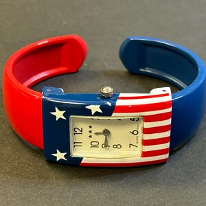 May include: A patriotic quartz watch with a red, white, and blue design. The watch face features the American flag with stars and stripes. The watch band is a cuff style, with red and blue sections.