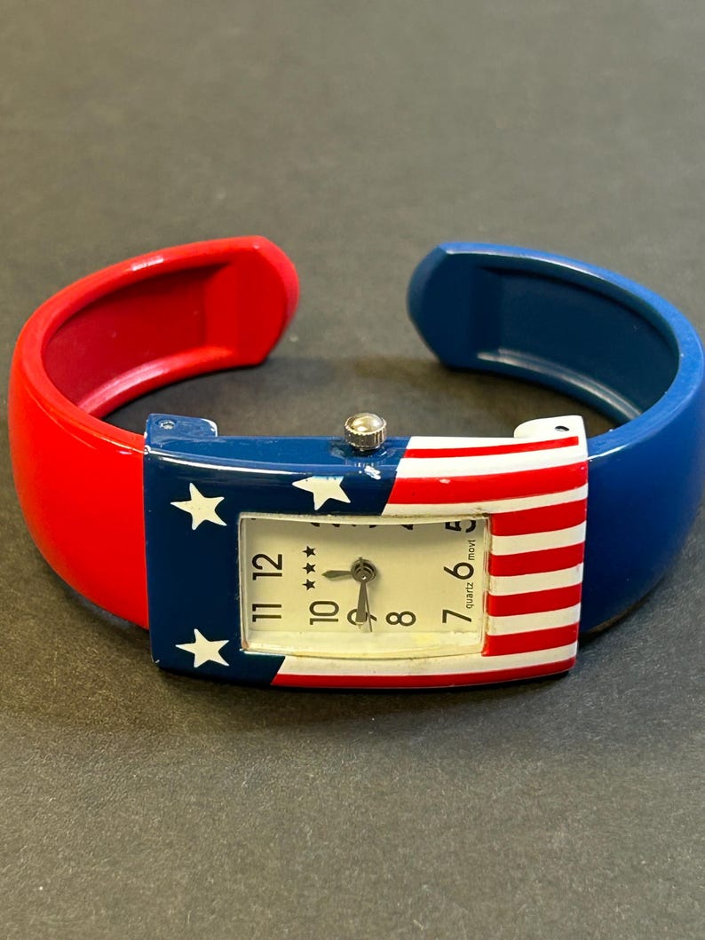 May include: A patriotic quartz watch with a red, white, and blue design. The watch face features the American flag with stars and stripes. The watch band is a cuff style, with red and blue sections.