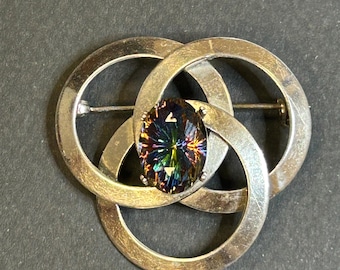Pode incluir: Um broche de prata com uma grande gema iridescente no centro. O broche tem a forma de três círculos entrelaçados.