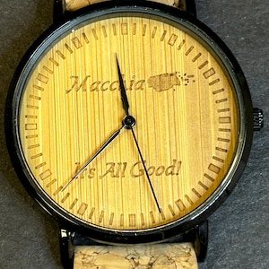 May include: A black and wood wristwatch with a cork band. The watch face has a wood background with the words "Macchia" and "It's All Good!" engraved on it.