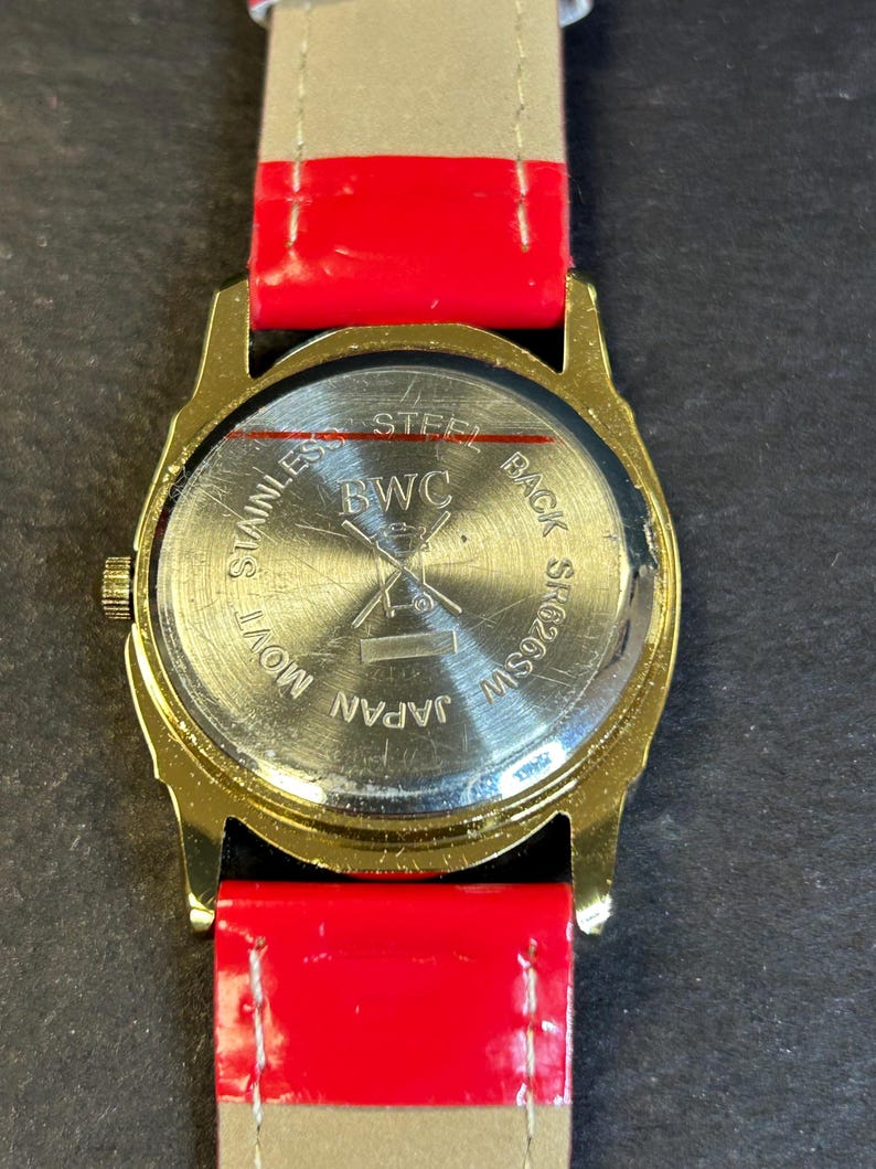May include: Gold-tone watch with a red and white striped leather band. The back of the watch has the text "Stainless Steel Back SS W259SW Japan Mov't BWC".